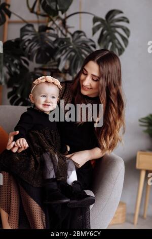 Beautiful mother sits on a chair and keeps her daughter on lap at home. family look concept. mother's, baby's day Stock Photo