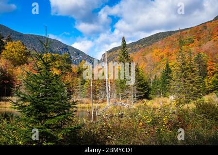 Majestic forested mountain scenery at the peak of fall foliage on a sunny day. A pond surrounded by colourful deciduous trees is in foreground. Stock Photo