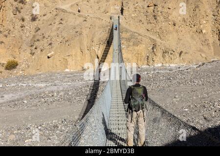 A man with a backpack stands on a suspension pedestrian bridge over a mountain gorge and looks at the mountains. Nepal. Himalayas. Stock Photo