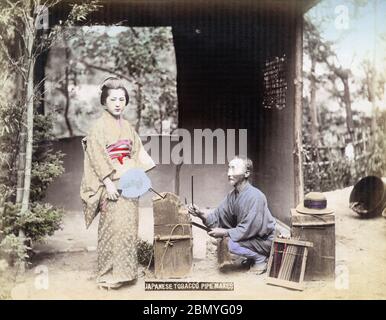 [ 1890s Japan - Japanese Pipe Mender ] —   A pipe mender wearing a happi coat works on a kiseru pipe (煙管) while a young woman poses.  19th century vintage albumen photograph. Stock Photo