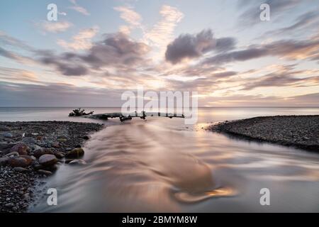 Picturesque sunset on a stone beach, view along a small river that flows into the sea, alluvial tree in front of the horizon, waves, long time exposur Stock Photo