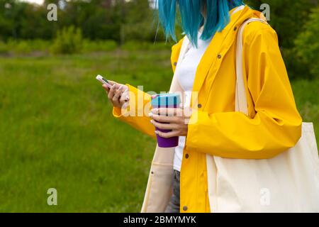 Young adult beautiful woman with blue hair standing in the outdoor garden holding a reusable cup with hot drink Stock Photo