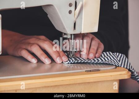 Woman hands using the sewing machine to sew a colorful face medical mask during the coronavirus pandemic. Homemade DIY protective mask against the virus. Sewing masks, COVID-19. Selective focus. Stock Photo