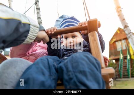 Two cute adorable playful caucasian siblings boy girl child enjoy having fun swinging wooden swing at backyard together with father. Little toddler Stock Photo