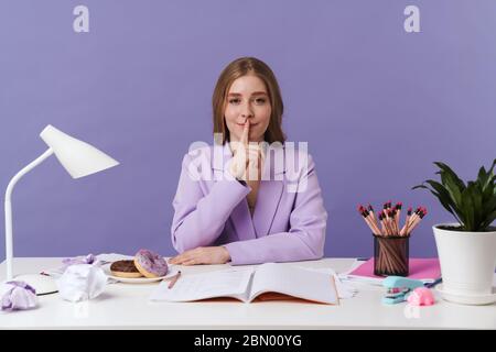 Photo of a positive optimistic young woman sit at the table indoors isolated over purple wall background showing silence gesture. Stock Photo