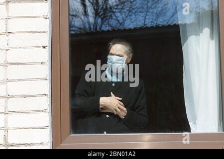 An old grandmother in a protective mask looks out the window at self-isolation. Stock Photo
