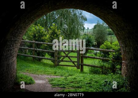 A rustic wooden country gate and fence, trees and bushes viewed through the end of a railway tunnel path during fine weather in summer Stock Photo