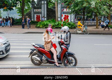 A young woman sitting on a motorbike and a young man standing each wearing face masks and helmets look at their mobile phones in Hanoi, north Vietnam Stock Photo