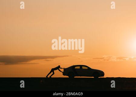 Silhouette of man driver pushing his car along on an empty road after breakdown at sunset, copy space, side view. Stock Photo