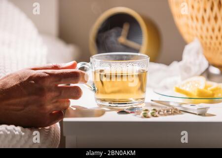 Sick man laying in bed under blanket and holding cup of hot tea with lemon, close up. Male drinking hot beverage to get well from flu, fever and virus Stock Photo