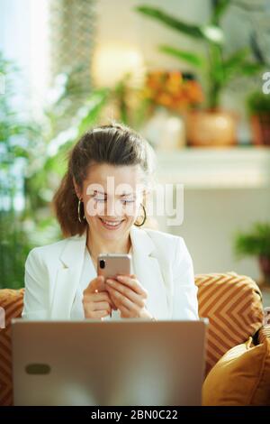 happy modern woman in white blouse and jacket with laptop and smartphone texting in the modern living room in sunny day. Stock Photo