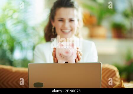 happy elegant middle age housewife in white blouse and jacket with laptop and piggy bank in the modern house in sunny day. Stock Photo