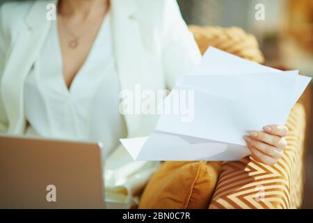 Closeup on woman with documents using laptop while sitting on divan in the house in sunny day. Stock Photo