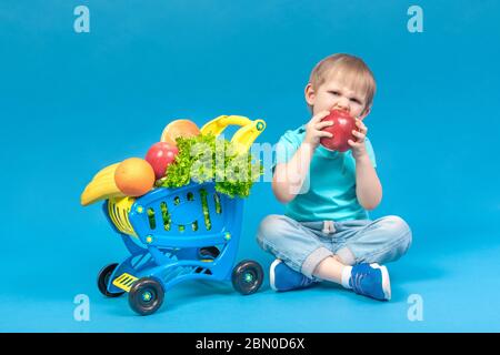 A hungry blond child sits near a supermarket basket full of food carts and bites a huge red apple. The concept of food delivery and healthy eating fru Stock Photo