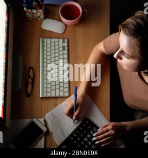 Woman using calculator to calculate the invoice, planning expenses, holding a pen, working on desktop pc late at night at home office, can not sleep.F Stock Photo