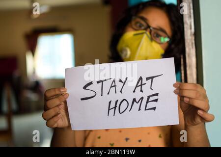 Little Indian girl wearing face mask holds a placard in hands showing a message 'Stay at Home' during COVID-19 pandemic to maintain social distancing.