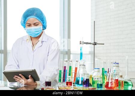 Young serious Asian female laboratory worker filling form on tablet computer after doing research in clinic Stock Photo