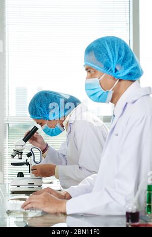 Laboratory technician looking through microscope when her colleague working on computer and filling research documents Stock Photo