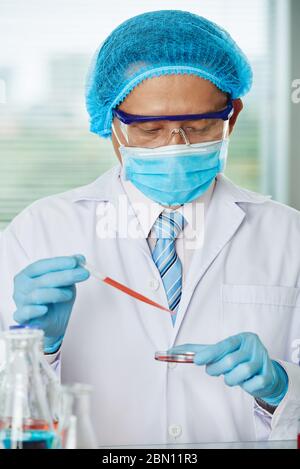 Serious Asian researcher in medical mask and protective glasses putting some liquid in petri dish Stock Photo