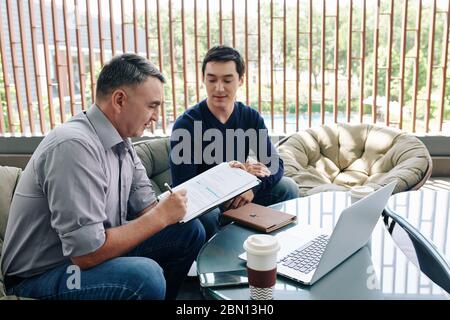 Mature entrepreneur signing contract with young software developer after having meeting in office lounge area Stock Photo