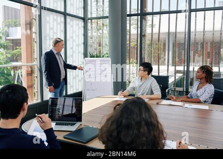 Experienced business trainer conducting effective training session for business team in meeting room and pointing at questions on white board Stock Photo