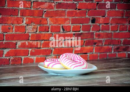 Two donuts in pink strawberry glaze on a white plate stand on a wooden table against a brick wall. Close-up. Stock Photo