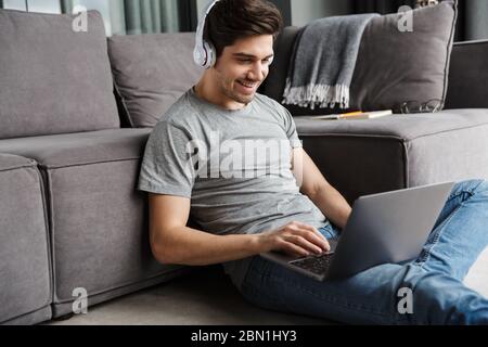 Portrait of an attractive smiling young bearded man wearing casual clothes sitting on a floor at the couch in the living room, using laptop computer, Stock Photo
