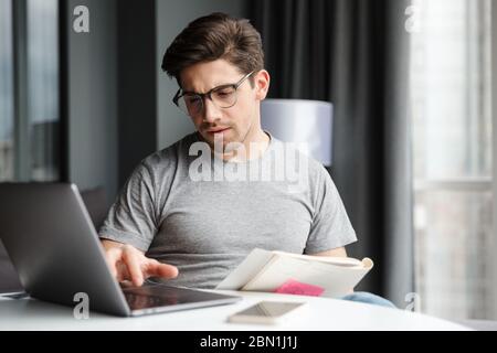 Handsome serious young bearded man wearing casual clothes using laptop computer while sitting at the table at home Stock Photo