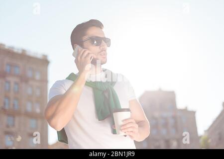 Young brunete man in a cozy outfit is talikg on the mobile, outdoors. He is on a walk outside, enjoying, holding a plastic cup of coffee in his hand Stock Photo