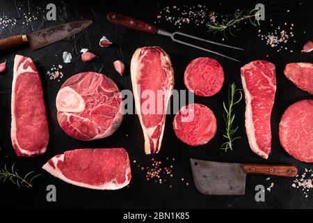 Cuts of meat, overhead flat lay shot on a dark background with salt, pepper, rosemary and knives Stock Photo