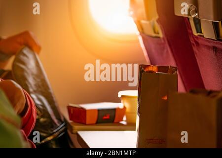 Food in brown paper bag put on plastic airplane tray table at seat back with blurred passenger hand opened black leather bag. Sunlight passing through Stock Photo