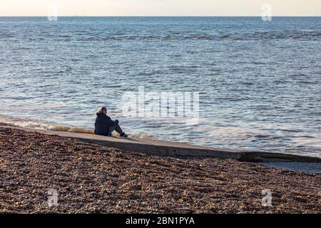 A woman sits on a slipway on the pebble beach at Hampton, Herne Bay, and stares out to sea, thoughtfully, UK Stock Photo