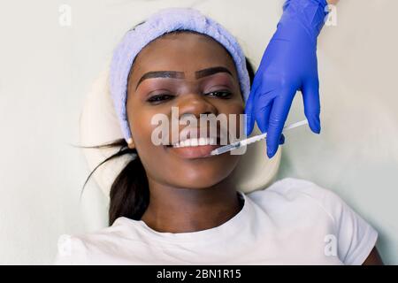 Beautiful young African woman gets cosmetic injection on her lips, isolated over light background. Doctor is making an injection in lips. Beauty Stock Photo