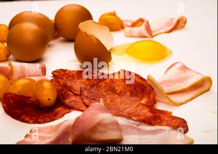 Broken chicken egg, scattered chicken eggs, slices of sausage and bacon and tomatoes on a white background. Close up Stock Photo