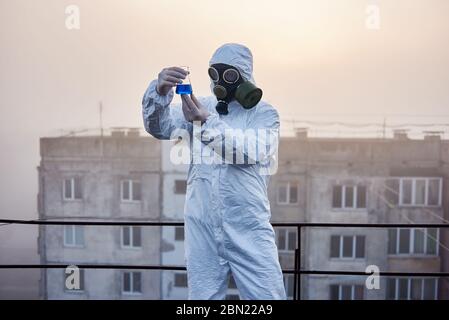 Scientist wearing white protective coverall, gloves and gas mask looking at a glass flask with blue reagent that he holds in his hands, block of flats on background Stock Photo