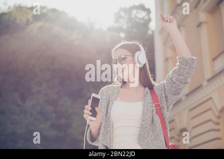 Funky spring mood! Chilling on the sun. Young happy lady is listening to the music outdoors on her phone, wearing sunglasses and big white earphones Stock Photo
