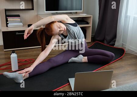 Red-haired girl is engaged in stretching at home in front of a laptop.