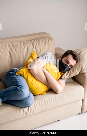 Nervous young man in cloth mask feeling stressed in quarantine embracing pillow on sofa and looking with locked eyes at one point Stock Photo