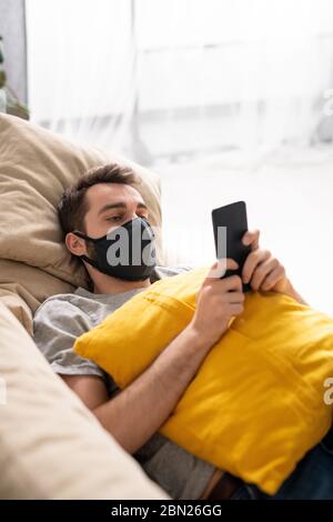 Despondent young man in black mask lying on sofa in living room and checking coronavirus statistics on phone Stock Photo
