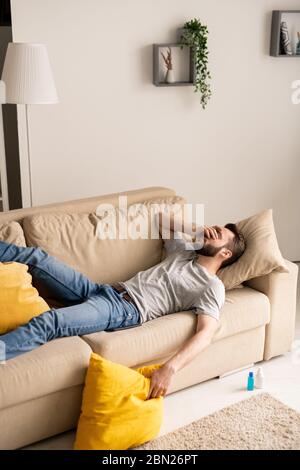 Young bearded man being in depression because of coronavirus isolation lying on sofa and covering face with hand Stock Photo