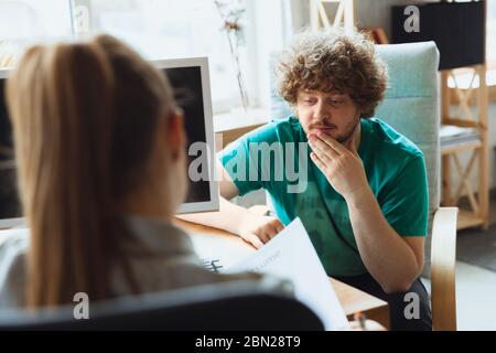 Young man sitting in office during the job interview with female employee, boss or HR-manager, talking, thinking, looks confident. Concept of work, getting job, business, finance, communication. Stock Photo