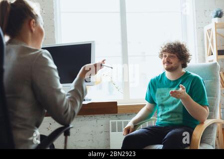 Young man sitting in office during the job interview with female employee, boss or HR-manager, talking, thinking, looks confident. Concept of work, getting job, business, finance, communication. Stock Photo
