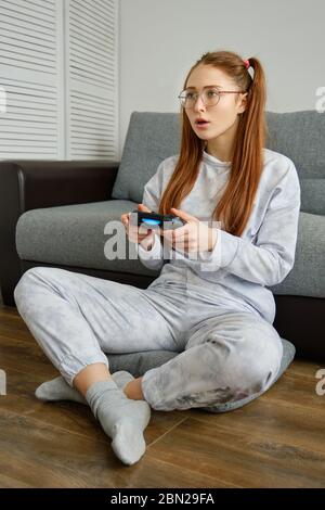Redhead girl in pajamas sits on the floor with a game joystick and looks in surprise in front of her Stock Photo
