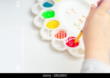 Closeup of hand of child holding brush over palette of watercolors on white sheet of paper. Stock Photo