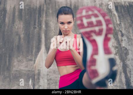 Self defence. Focus of a young cute serious fighter, training kickboxing high kick exercise with her foot, outdoors, in pink fashionable sport outfit