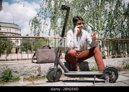 Attractive guy sitting on curb and talking on the phone. Brunette man with sunglasses speaking on the phone while sitting on curb next to scooter. Stock Photo