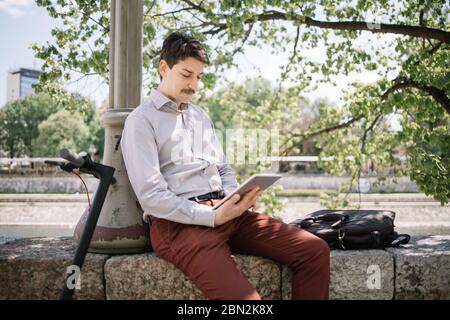 Young man holding tablet while sitting on fence and leaning on pillar. Male freelancer using tablet while sitting on stone fence under green tree. Stock Photo