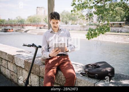 Serious man sitting on stone fence and looking at tablet. Freelancer man using tablet while sitting on stone fence next to scooter with river in backg Stock Photo