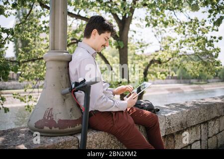Side view of guy with tablet sitting on fence with tree shade. Man using tablet and leaning on pillar in front of city river under green tree. Stock Photo