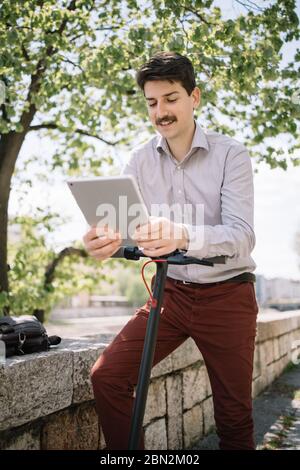 Portrait of man with tablet leaning on scooter rudder. Young man propping himself up on scooter rudder while using tablet and standing under tree near Stock Photo